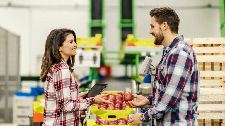 Duas pessoas sorrindo e interagindo em um armazém de alimentos, uma segurando uma maçã e a outra usando um dispositivo móvel de digitalização.