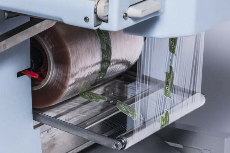 Detalhe da máquina Elixa 21 da Teixpac em funcionamento, mostrando filme plástico sendo enrolado com etiquetas da marca 'Orgânico'.