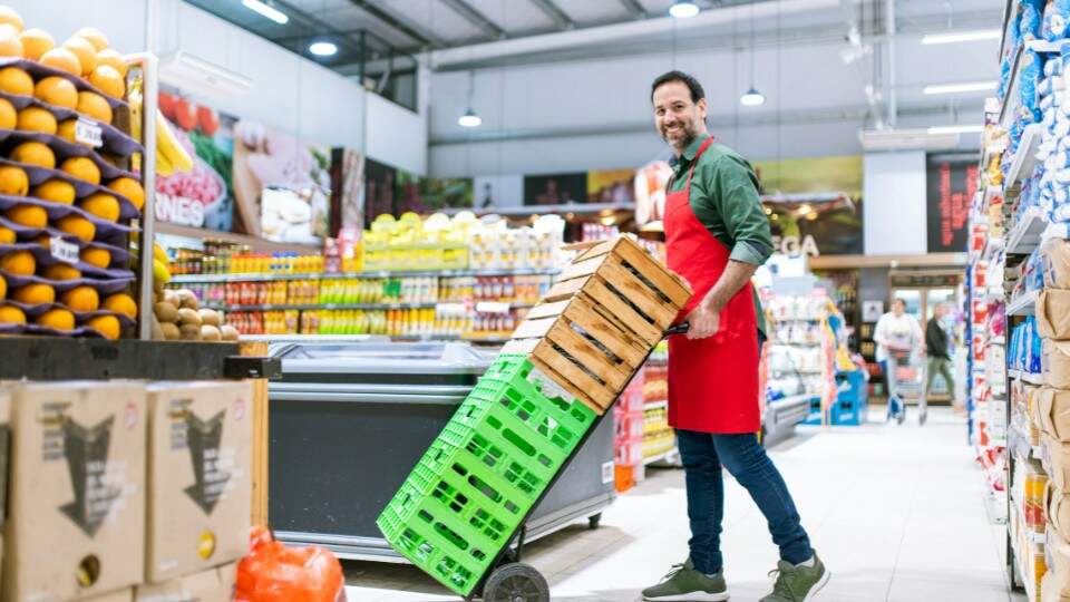 Funcionário de supermercado sorridente, vestindo um avental vermelho, empurrando um carrinho com caixas verdes em um corredor cheio de produtos.